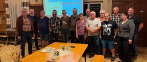 Eine Gruppe von Menschen steht vor einer Projektionsleinwand, lächelt und posiert für ein Foto. Der Tisch hat Papiere, ein Buch und ein Glas. Der Raum hat Holzböden und eine Gardine auf der linken Seite.