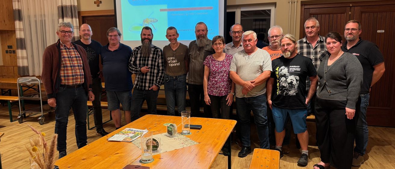 Eine Gruppe von Menschen steht vor einer Projektionsleinwand, lächelt und posiert für ein Foto. Der Tisch hat Papiere, ein Buch und ein Glas. Der Raum hat Holzböden und eine Gardine auf der linken Seite.