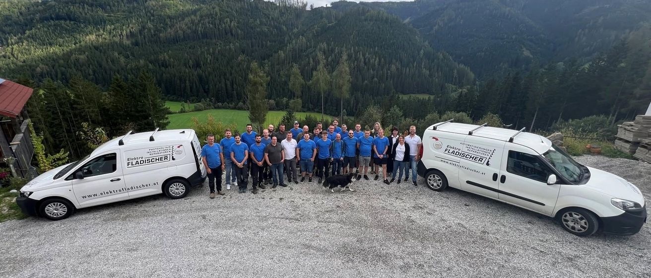 Eine Gruppe von Menschen in blauen Hemden und Mützen steht vor zwei weißen Lieferwagen mit Text auf einem Schotterweg, mit Bergen und Grün im Hintergrund.
