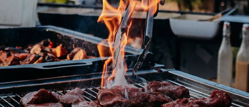 Ein Koch hält Zangen über einem Grill mit Flammen. Der Grill hat Essen und Flaschen in der Nähe. Bäume und Gebäude sind im Hintergrund.