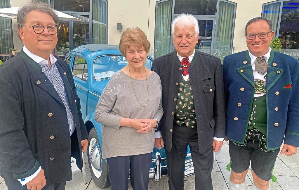 Vier Personen, zwei Männer und zwei Frauen, stehen vor einem blauen Oldtimer. Sie lächeln und scheinen für ein Foto zu posieren. Der Mann auf der rechten Seite trägt ein traditionelles bayerisches Outfit.