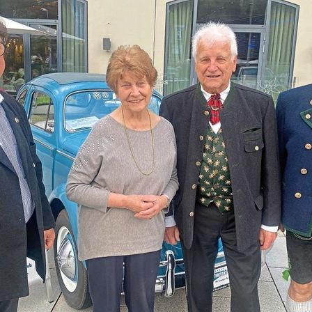 Vier Personen, zwei Männer und zwei Frauen, stehen vor einem blauen Oldtimer. Sie lächeln und scheinen für ein Foto zu posieren. Der Mann auf der rechten Seite trägt ein traditionelles bayerisches Outfit.