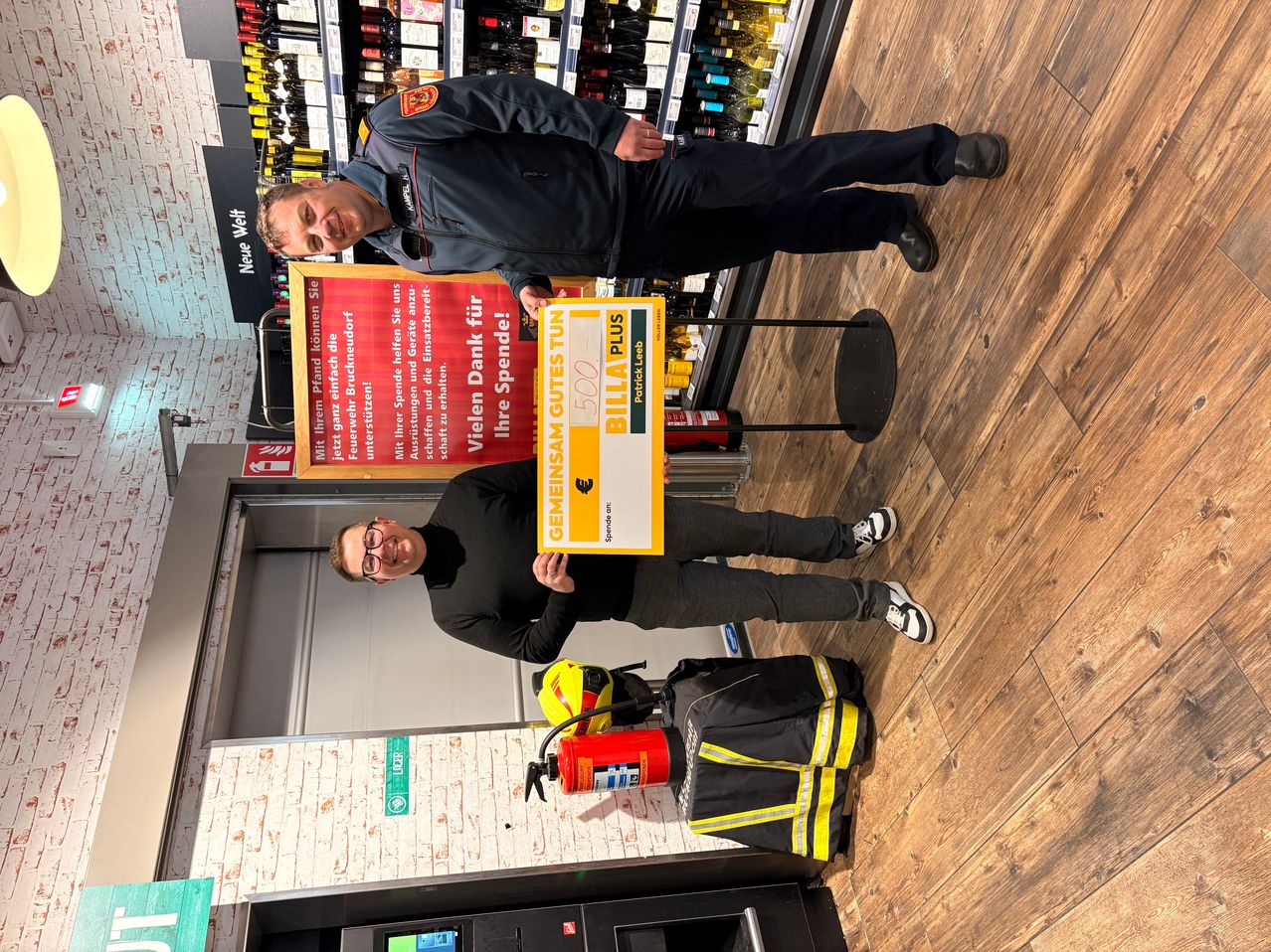 Zwei Männer stehen in einem Geschäft mit einem Schild. Ein Mann hält ein Schild mit der Aufschrift Gemeinsam Gutes tun und einem Scheck. Dahinter steht ein Regal mit Flaschen und einem Feuerlöscher.