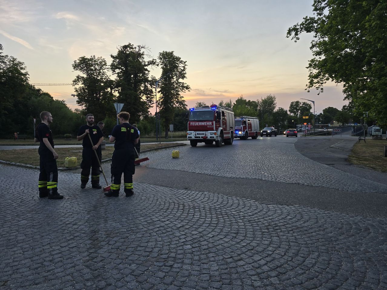 Firefighters are cleaning up a spill on a cobblestone road as fire trucks drive by.