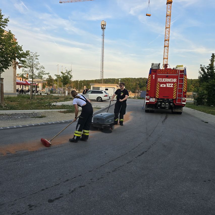 Two firefighters clean the road with a street sweeper while a fire truck is parked nearby.