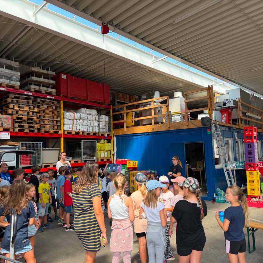 Eine Gruppe von Kindern und Erwachsenen versammelt sich in einem Lagerhaus, mit Regalen im Hintergrund, die mit verschiedenen Artikeln gefüllt sind. Eine Frau steht vor ihnen und gibt möglicherweise Anweisungen.