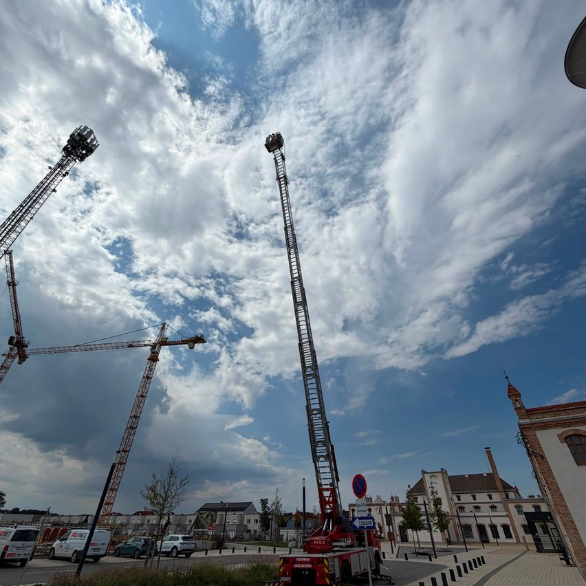 Ein Baugelände mit zwei Kränen und einem Feuerwehrauto unter einem bewölkten Himmel. Im Hintergrund sind Gebäude und geparkte Autos zu sehen.