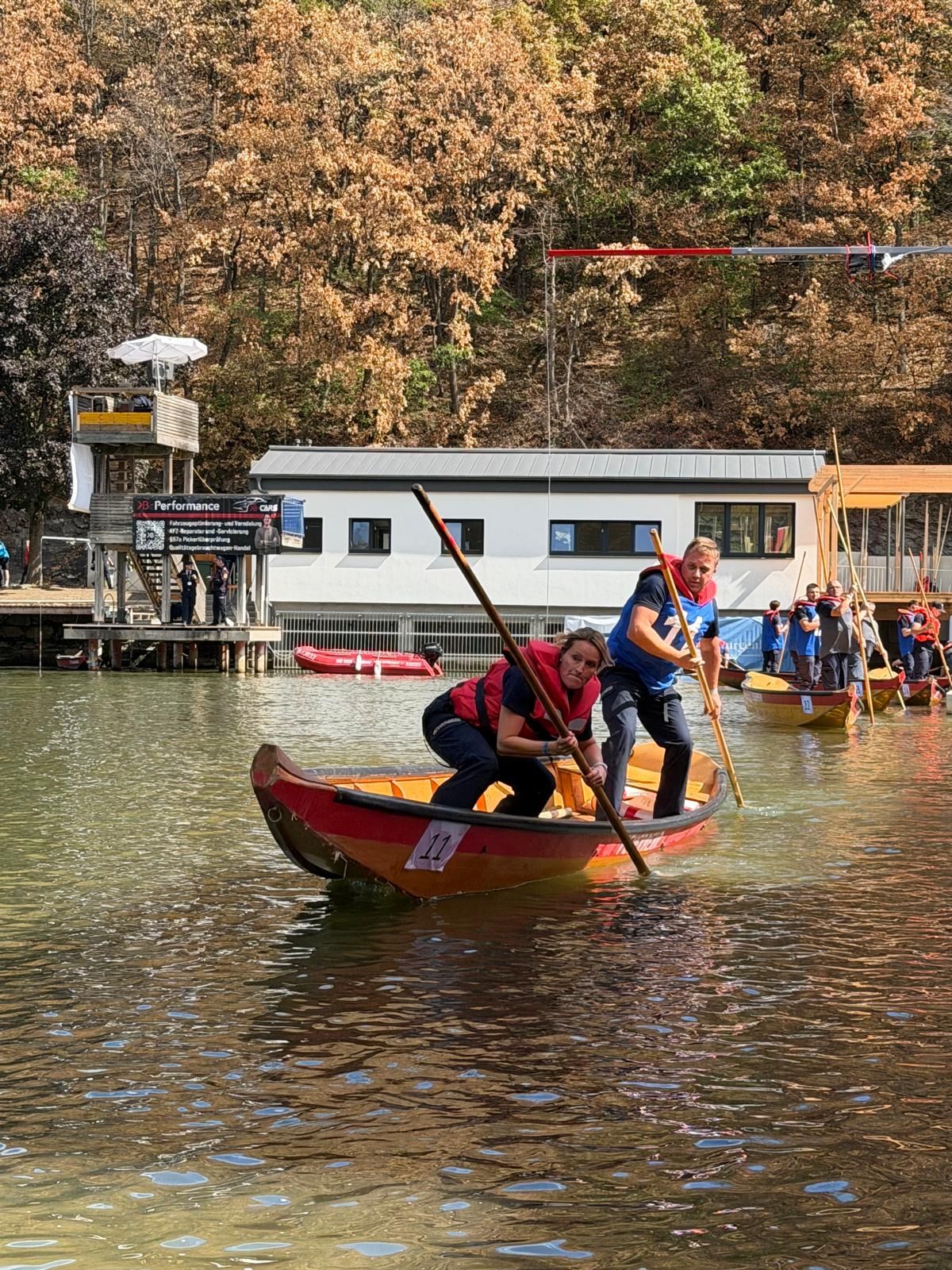 Zwei Personen in einem roten Boot paddeln auf einem Fluss, mit einem Steg und einem Gebäude im Hintergrund.