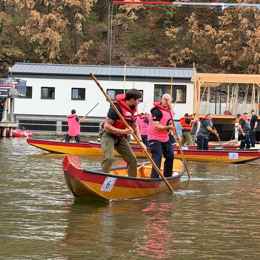 Eine Gruppe von Menschen in Rettungswesten paddelt auf einem Fluss, mit einem Gebäude und Bäumen im Hintergrund.