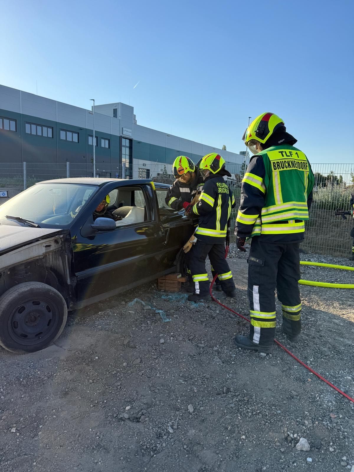 Rettungskräfte in Schutzausrüstung arbeiten daran, eine Person aus einem beschädigten Auto zu entfernen, mit einem Zaun und einem Gebäude im Hintergrund.