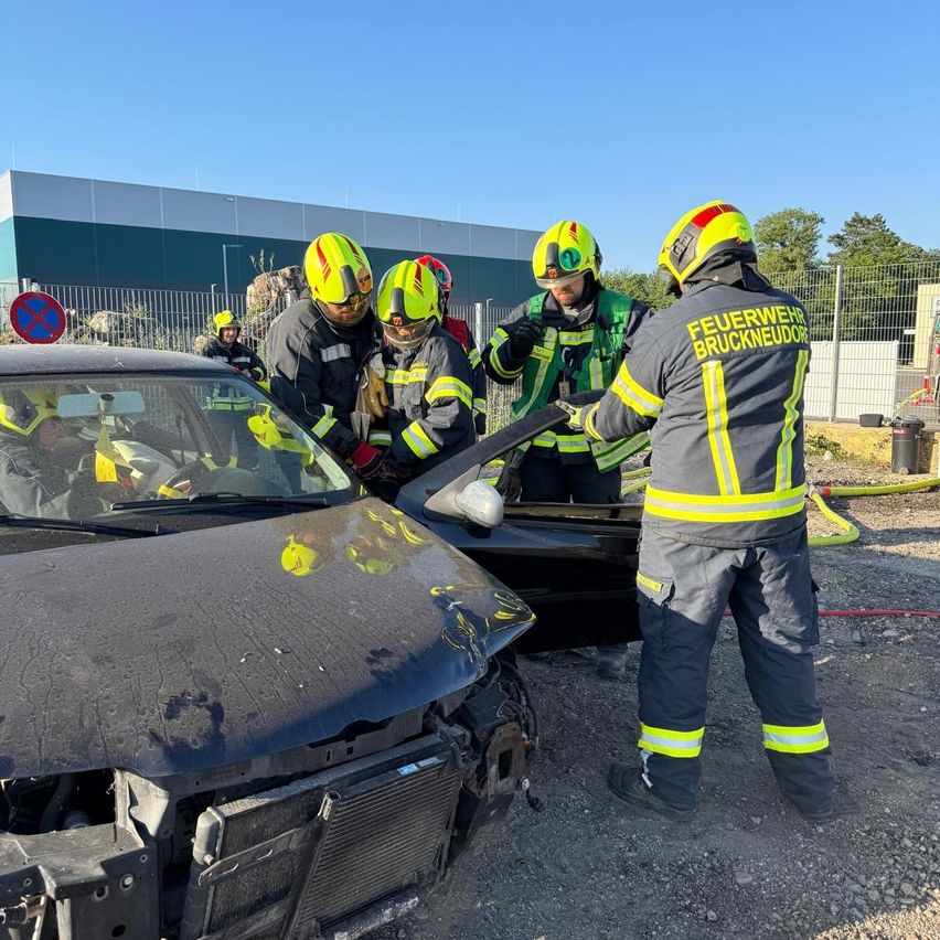 Eine Gruppe Feuerwehrleute in voller Ausrüstung inspiziert ein demoliertes Auto. Ein Feuerwehrmann hält ein Werkzeug in der Nähe des Stoßfängers des Autos.