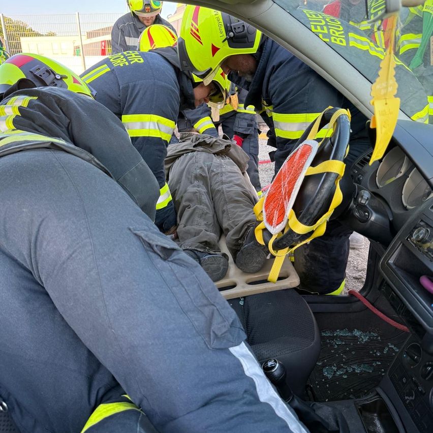 Feuerwehrleute in gelben Helmen und Uniformen retten eine Person aus einem zerstörten Auto. Ein Feuerwehrmann sitzt auf dem Beifahrersitz, während ein anderer eine Trage hält.