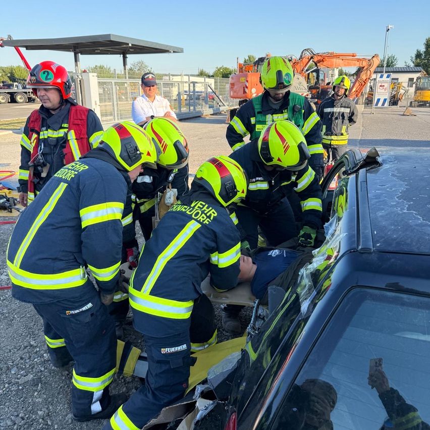 Feuerwehrleute in gelben und schwarzen Uniformen mit Helmen retten jemanden aus einem beschädigten Auto. Ein erwachsener Mann beobachtet sie.