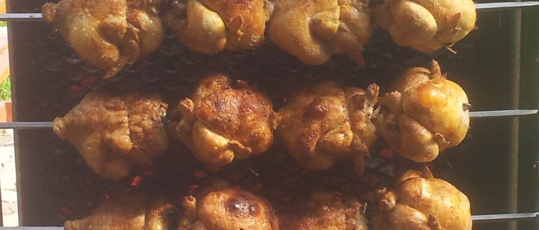 Ein Nahaufnahme eines Grills mit mehreren Spießen von ganzen Hähnchen beim Garen. Die Hähnchen haben eine knusprige, goldbraune Haut. Der Hintergrund ist leicht verschwommen.