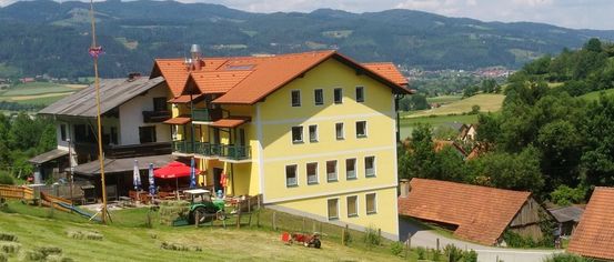 A yellow house with a red roof, multiple windows, and balconies sits on a grassy hillside with hay bales. A tractor and machinery are parked nearby.