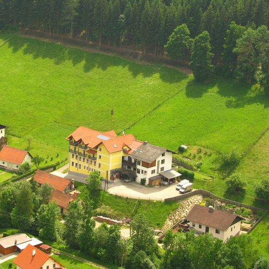 Luftaufnahme eines kleinen Dorfes mit mehreren Häusern und Gebäuden, umgeben von grünen Feldern und Bäumen. Ein gelbes und weißes Gebäude mit rotem Dach steht im Zentrum.