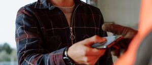 Ein lächelnder Mann in einem karierten Hemd hält ein Handy in der Hand und führt wahrscheinlich eine Zahlung durch. Er steht vor einer weißen Wand mit einem Ausfahrtsschild.