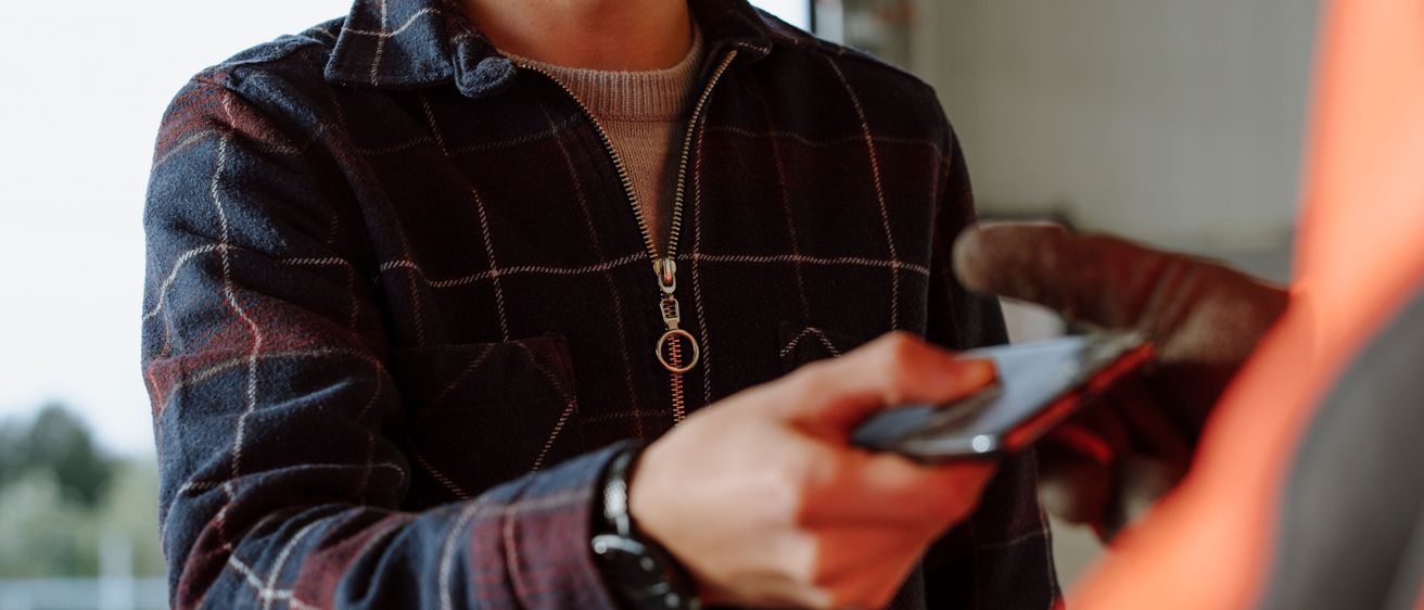 Ein lächelnder Mann in einem karierten Hemd hält ein Handy in der Hand und führt wahrscheinlich eine Zahlung durch. Er steht vor einer weißen Wand mit einem Ausfahrtsschild.