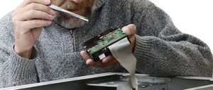 Ein Mann mit Brille und grauem Pullover arbeitet an einem Computer. Er hält einen Schraubendreher und eine Leiterplatte.