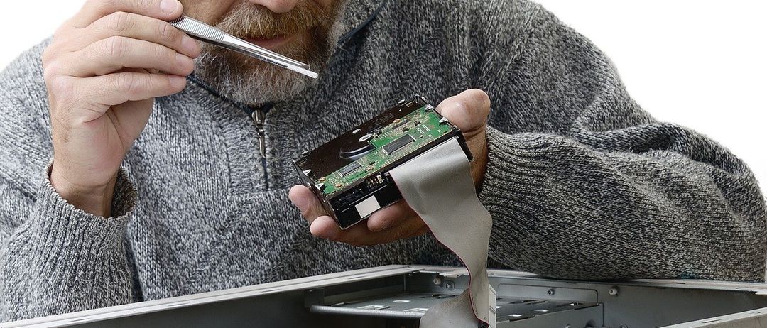 Ein Mann mit Brille und grauem Pullover arbeitet an einem Computer. Er hält einen Schraubendreher und eine Leiterplatte.