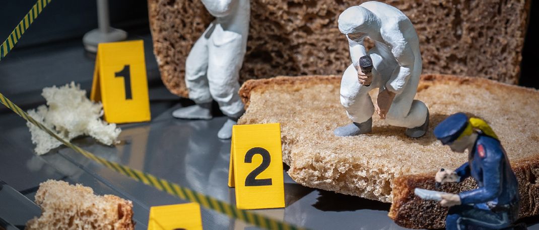 Ein Kriminalszenen-Tableau mit drei Figuren auf einem Tisch. Eine in einem Anzug fotografiert, eine andere in einem Anzug schreibt und eine dritte in Uniform inspiziert ein Stück Brot.