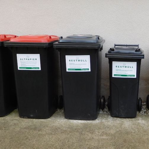 Fünf schwarze Behälter mit verschiedenfarbigen Deckeln sind an einer Wand aufgestellt. Die Behälter haben weiße Etiketten mit Text und Logos.