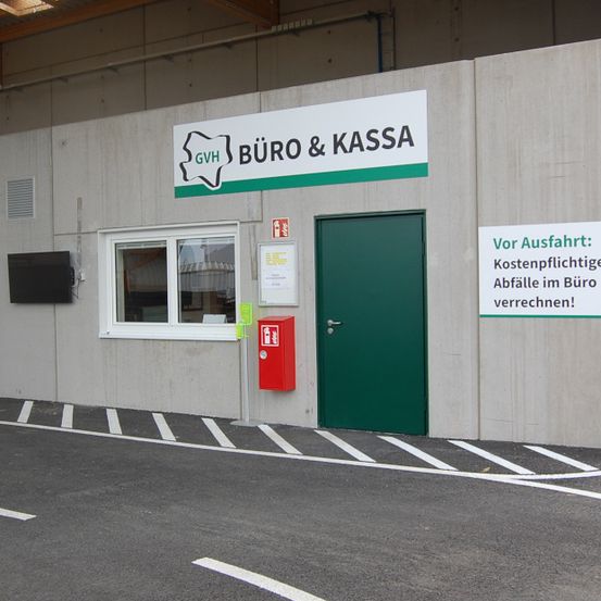 Der Eingang zu einem Gebäude mit der Aufschrift 'Büro & Kassa' und einer grünen Tür. Ein Schild weist darauf hin, dass 'Vor Abfahrt: Kostenpflichtige Abfälle im Büro verrechnet!' werden. Ein Monitor ist an der Wand montiert.