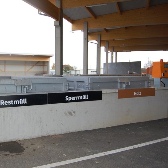 Eine Betonbarriere mit zwei Schildern: eines steht 'Restmuell' und das andere 'Sperrmueell'. Hinter der Barriere befindet sich ein Gebäude mit einer Holzkonstruktion. Der Himmel ist bedeckt.