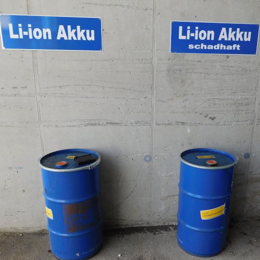 Drei blaue Fässer stehen am Boden in der Nähe einer Wand. Zwei blaue Schilder über den Fässern lesen Li-ion Akku und Li-ion Akku schadhaft.