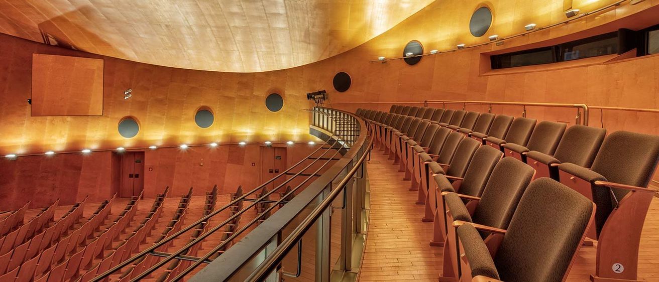 Ein leerer Konzertsaal mit modernem Design, gebogenen Sitzreihen, Holzböden und kreisförmigen Beleuchtungskörpern.