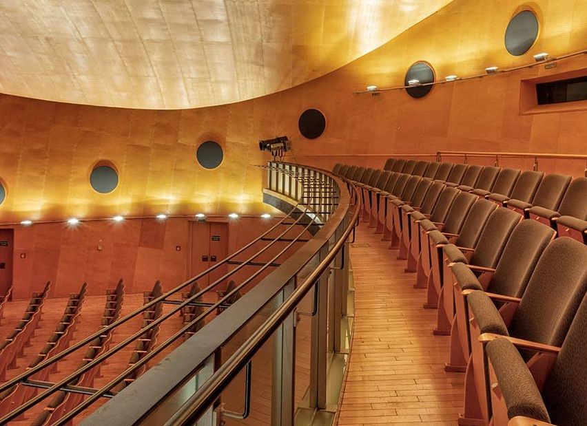 Ein leerer Konzertsaal mit modernem Design, gebogenen Sitzreihen, Holzböden und kreisförmigen Beleuchtungskörpern.