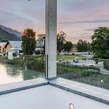 Ein Balkonausblick mit Fluss, Bergen und einer Stadt bei Sonnenuntergang. Bäume säumen den Fluss und mehrere Autos sind in der Nähe geparkt.