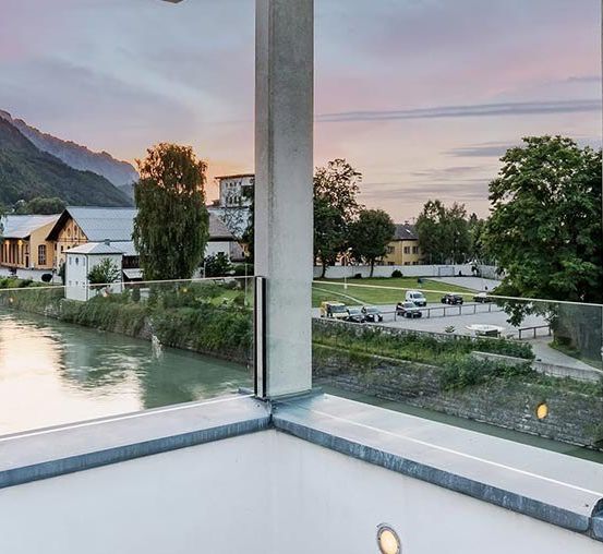 Ein Balkonausblick mit Fluss, Bergen und einer Stadt bei Sonnenuntergang. Bäume säumen den Fluss und mehrere Autos sind in der Nähe geparkt.
