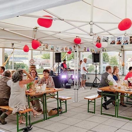Eine Gruppe von Menschen sitzt unter einem Zelt mit einem DJ in der Ecke. Das Zelt ist mit roten und weißen Luftballons und Fotos dekoriert.