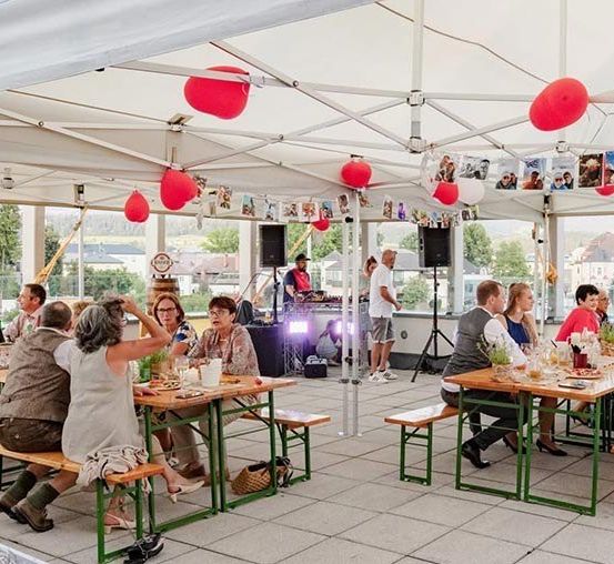 Eine Gruppe von Menschen sitzt unter einem Zelt mit einem DJ in der Ecke. Das Zelt ist mit roten und weißen Luftballons und Fotos dekoriert.