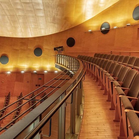Ein leeres Theater mit braunen Stühlen und einer goldenen Wand mit runden Fenstern. Die Galerie hat ein Metallgeländer.