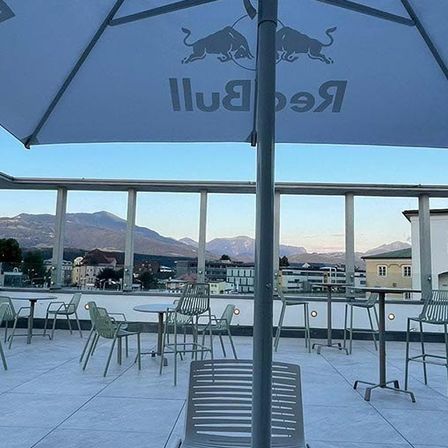 Eine Außensitzfläche mit Tischen und Stühlen, von denen einer einen Red Bull-Schirm hat. Dahinter sind Berge und Gebäude zu sehen.