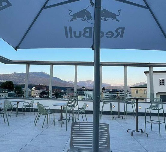 Eine Außensitzfläche mit Tischen und Stühlen, von denen einer einen Red Bull-Schirm hat. Dahinter sind Berge und Gebäude zu sehen.