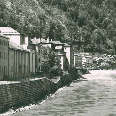 Ein altes Foto zeigt eine Stadt am Fluss mit Gebäuden, einer Brücke und einem Berg im Hintergrund.