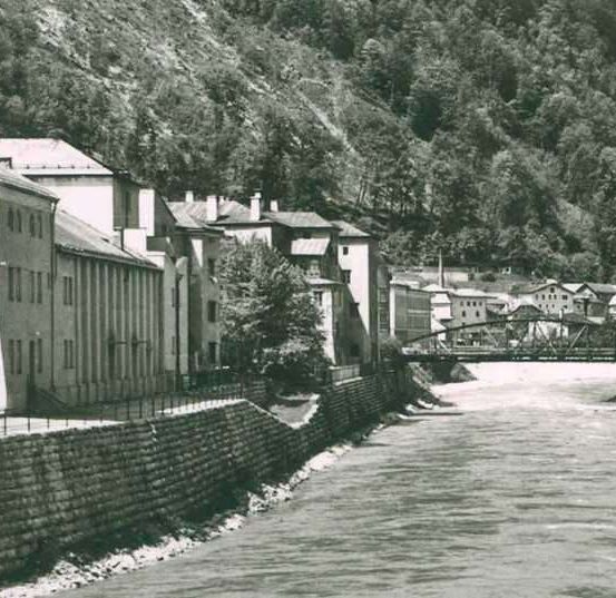 Ein altes Foto zeigt eine Stadt am Fluss mit Gebäuden, einer Brücke und einem Berg im Hintergrund.