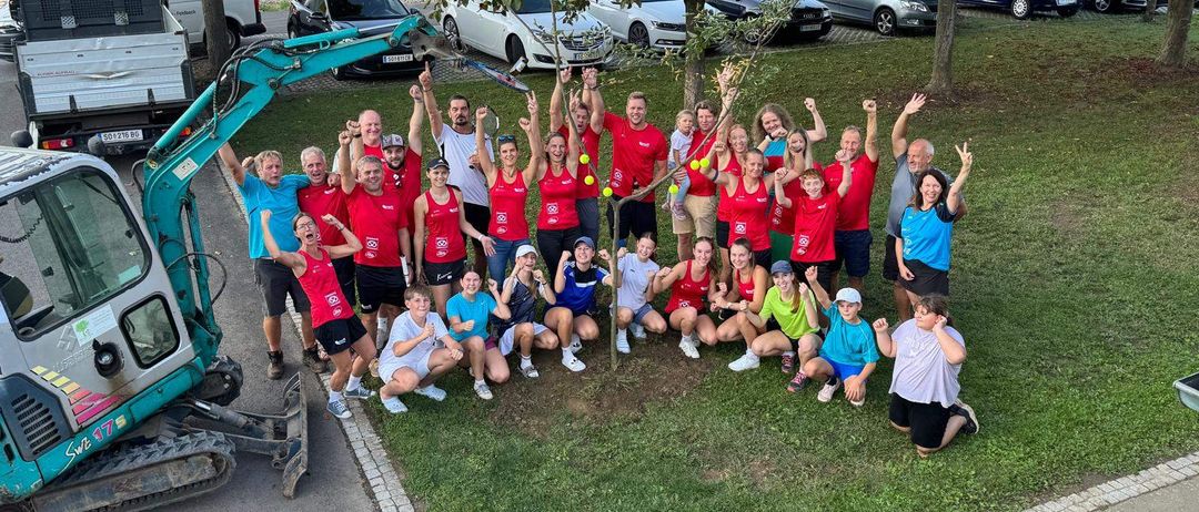 Eine Gruppe von Menschen in roten Shirts posiert für ein Foto um einen Baum, in der Nähe ein Baufahrzeug. Autos sind am Straßenrand geparkt.