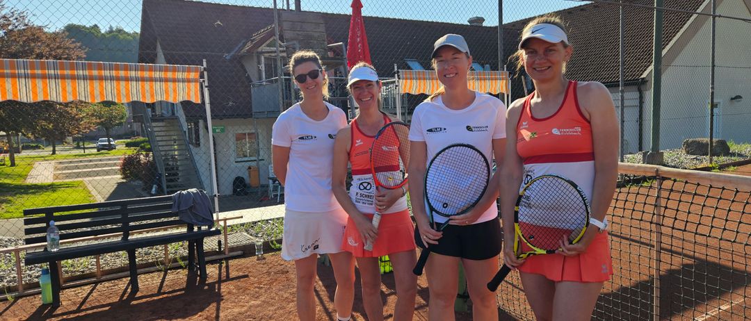 Vier Frauen stehen auf einem Tennisplatz und lächeln. Sie halten Tennisschläger in der Hand und tragen weiße und orangefarbene Uniformen. Hinter ihnen steht ein Gebäude mit einem Zaun und einem roten Schirm.