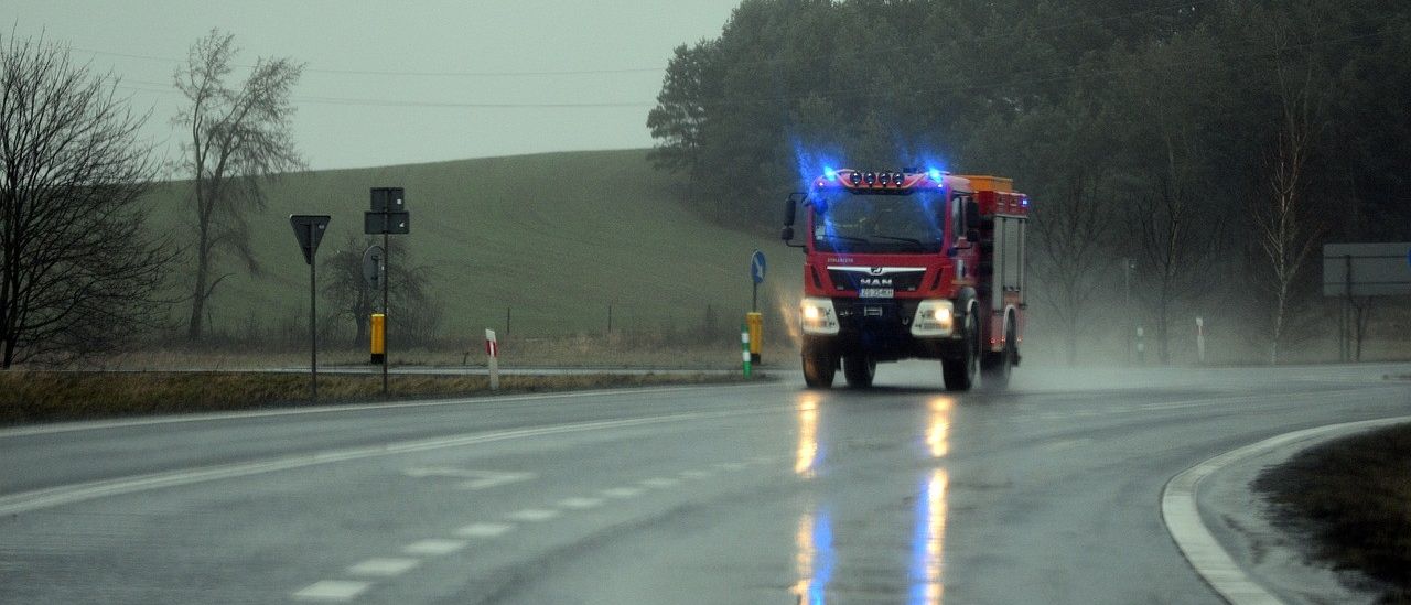 Ein roter Feuerwehrwagen mit blauen Blitzlichtern fährt auf einer nassen Straße, möglicherweise im Einsatz.