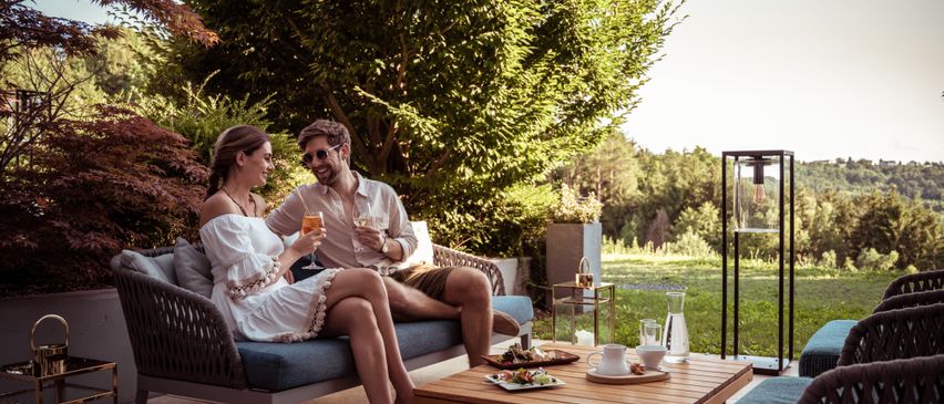 Ein Paar sitzt auf einer blauen Couch mit einem Holztisch davor. Sie lächeln und trinken aus Gläsern. Der Tisch hat Essen, eine Karaffe und Tassen.