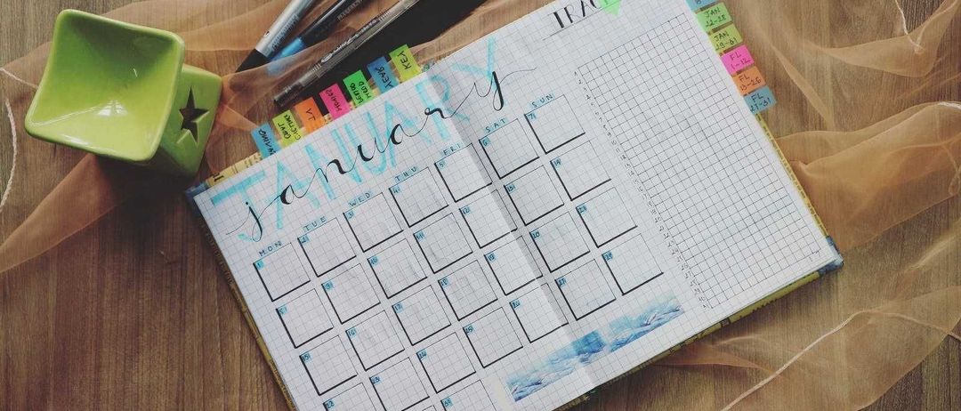 Ein geöffneter Planer mit dem hervorgehobenen Monat Januar liegt auf einer Holzoberfläche. Der Planer hat ein Rasterlayout mit farbigen Haftnotizen, die daran befestigt sind. Auf der linken Seite des Planers befinden sich Stifte, Marker und ein sternförmiges Objekt.