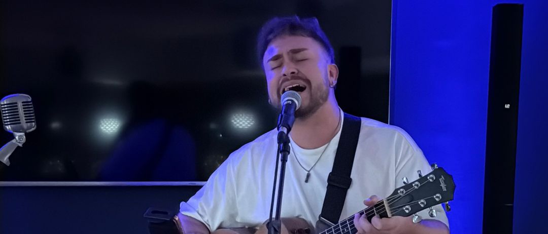 Ein Mann singt leidenschaftlich in ein Mikrofon, während er eine akustische Gitarre in einem blau beleuchteten Raum spielt.