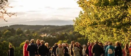 Eine Gruppe von Menschen und ein Hund stehen in einer Reihe auf einem Pfad, möglicherweise warten sie auf etwas. Sie tragen Hüte und einige haben Taschen dabei. Der Pfad ist von Gras und Bäumen umgeben. In der Ferne befinden sich Berge und ein klarer Himmel.
