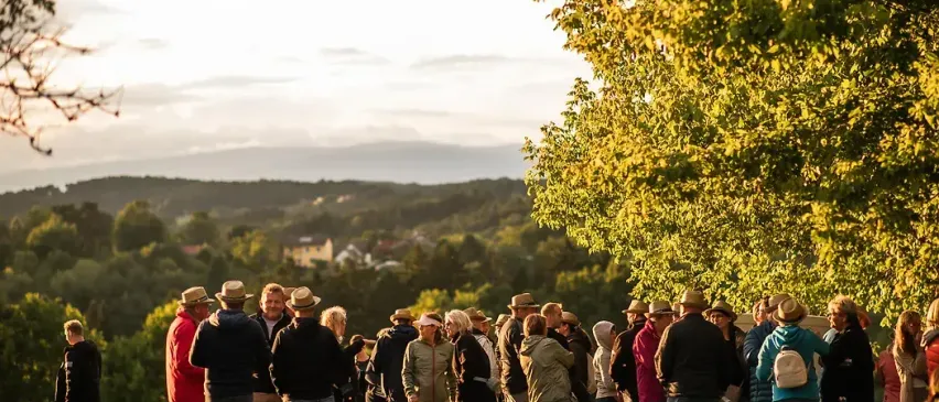 Eine Gruppe von Menschen und ein Hund stehen in einer Reihe auf einem Pfad, möglicherweise warten sie auf etwas. Sie tragen Hüte und einige haben Taschen dabei. Der Pfad ist von Gras und Bäumen umgeben. In der Ferne befinden sich Berge und ein klarer Himmel.
