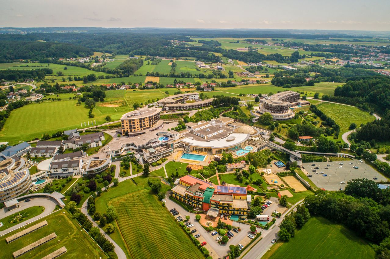 Luftaufnahme eines Hotelkomplexes mit mehreren Gebäuden, darunter ein großes kreisförmiges Gebäude mit Swimmingpool und mehrere Pools und einem Parkplatz im Vordergrund.