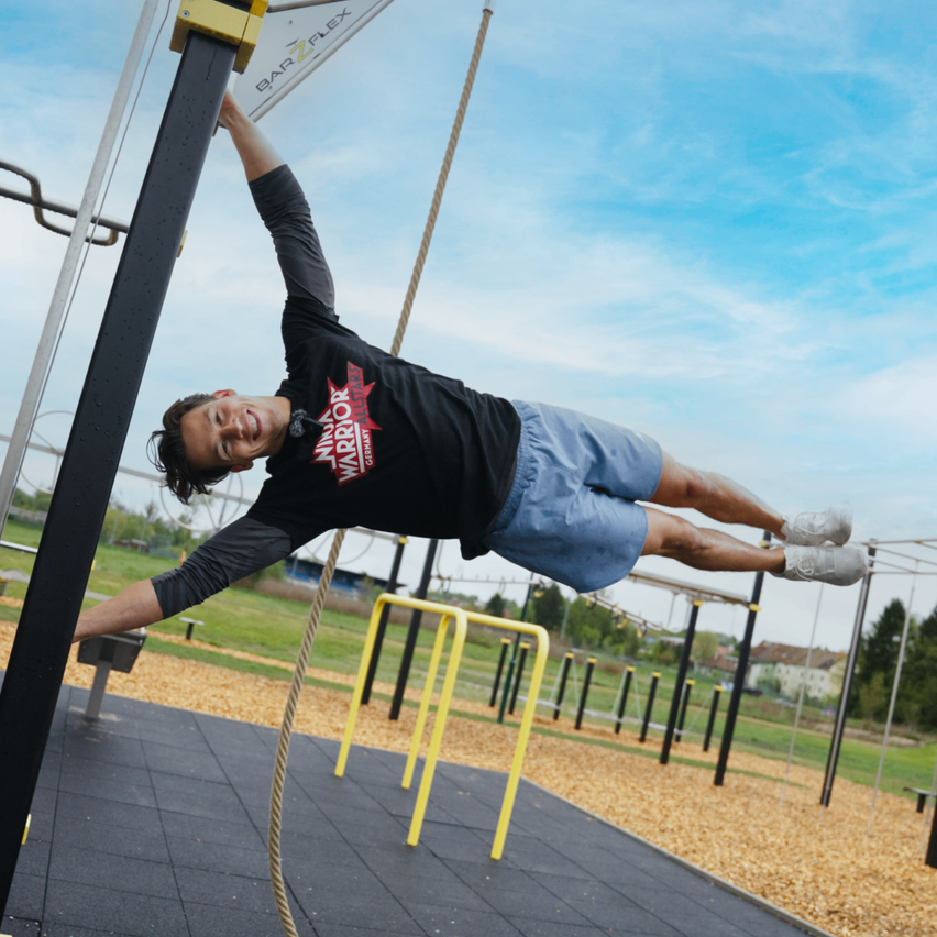 Ein Mann hängt kopfüber von einem Seil in einem Outdoor-Fitnessstudio, trägt ein schwarzes T-Shirt und blaue Shorts.